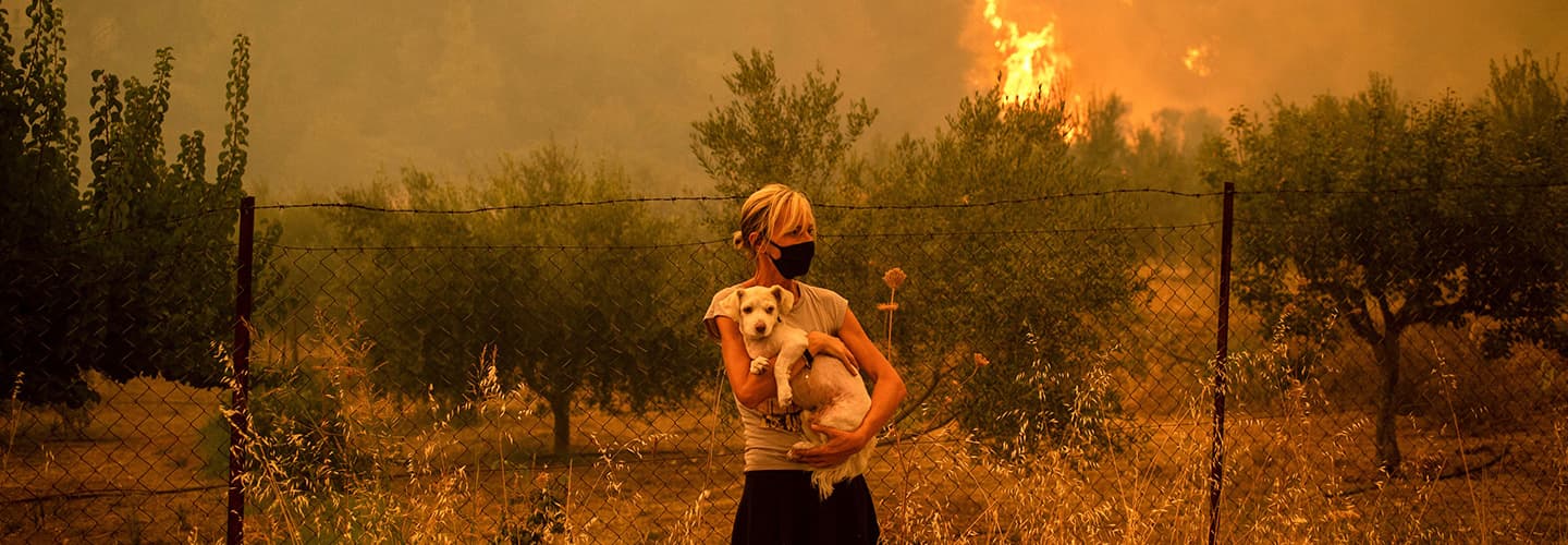 A woman holding a dog as wild fires blaze behind her